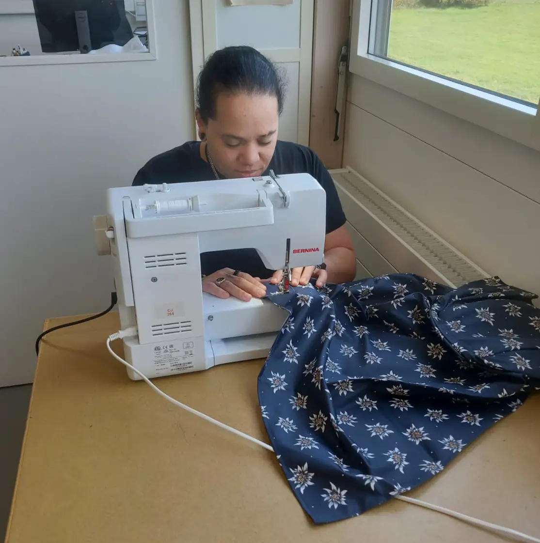 Mitarbeiterin näht einen Schürzenstoff mit Edelweiss-Muster an einer BERNINA-Nähmaschine in einem hellen Atelier mit Blick ins Grüne.