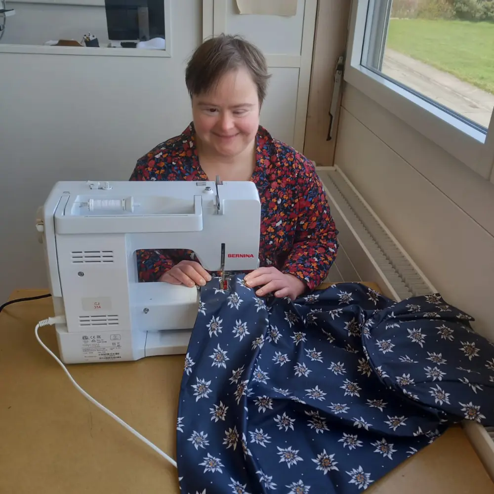 Lächelnde Mitarbeiterin näht einen Edelweiss-Schürzenstoff an einer BERNINA-Nähmaschine in einem geschützten Schweizer Atelier mit Tageslicht.